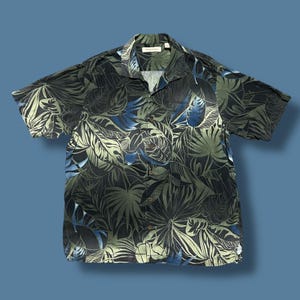 以下が含まれることがあります： 緑、青、黒の色合いの熱帯の葉模様の半袖ボタンダウンシャツ。 シャツは襟付きで、胸ポケットが1つ付いています。 生地は軽量に見えます。