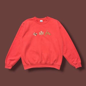 May include: A coral-colored sweatshirt with embroidered leaf designs. The crew neck sweatshirt features three embroidered leaves in shades of gold, brown, and green. The Gildan brand label is visible on the collar.