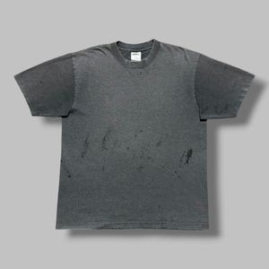 Pode incluir: Uma t-shirt de manga curta em cinzento-carvão com decote redondo e um aspeto desbotado e desgastado, apresentando manchas escuras dispersas e pequenos buracos por todo o tecido. A peça inclui uma etiqueta interior branca no pescoço com texto preto.