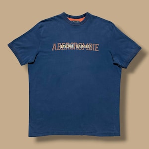 May include: A navy blue t-shirt with the text "Abercrombie Baseball Team 1892" in a rust-coloured font. The shirt has a crew neck with an orange interior and short sleeves. The shirt is made of a soft material.