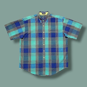 Op de afbeelding: Een shirt met korte mouwen in een blauw en turquoise ruitpatroon. Het shirt heeft een button-down kraag en een gele voering op de kraag. De stof is waarschijnlijk katoen. Het shirt wordt plat gepresenteerd tegen een effen achtergrond.