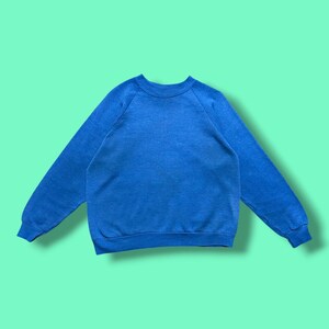 May include: A solid blue long-sleeve sweatshirt featuring a crew neckline, raglan sleeves, and ribbed cuffs at the wrists and hem. The garment is constructed from a plain, textured fabric in a vibrant shade of blue with no patterns or graphics.