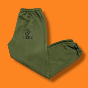 May include: Olive green sweatpants with the text "USMC" and a black emblem. The sweatpants have an elastic waistband and cuffs. The background is orange.