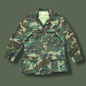 Può includere: Giacca mimetica nei toni del verde, marrone e beige. La giacca ha un collo, diverse tasche frontali e maniche lunghe. La giacca in stile militare è appoggiata su uno sfondo verde.
