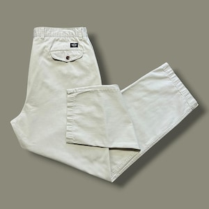 Puede incluir: Pantalones de algodón beige con cintura alta y trabillas para cinturón, un bolsillo trasero con solapa y cierre de botón, y una pequeña etiqueta rectangular de la marca sobre el bolsillo. Corte recto con costuras visibles.