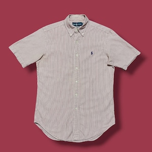 May include: A short-sleeved button-down shirt with vertical stripes in shades of red and white. The shirt features a button-down collar and a small blue logo on the chest. The shirt is made of a lightweight fabric.