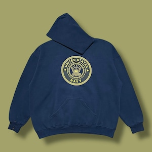 Könnte beinhalten: Marineblauer Kapuzenpullover mit Kängurutasche. Auf der Vorderseite befindet sich ein goldfarbenes rundes Emblem mit der Aufschrift "United States Navy". Das Emblem zeigt einen Adler und militärische Insignien.