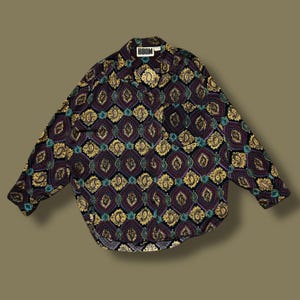 Pode incluir: Uma camisa de manga comprida com um padrão paisley geométrico em tons de roxo, dourado, verde-azulado e preto. A camisa tem gola e bolso. A palavra "BOOM" está impressa na etiqueta.
