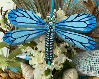 May include: A blue and brown wooden dragonfly with glitter accents hangs from a blue string. The dragonfly is surrounded by a variety of natural elements, including seashells, straw, and greenery.