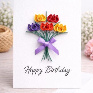May include: A white birthday card featuring a paper flower bouquet in shades of yellow, red, orange, and purple, tied with a purple ribbon. The card reads "Happy Birthday" in cursive script.