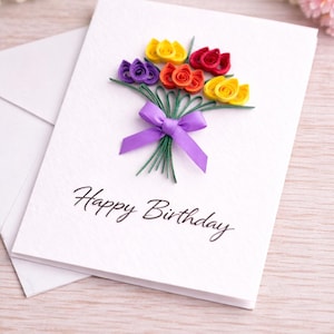 May include: A white birthday card featuring a paper flower bouquet in purple, yellow, red, and orange. The card is tied with a purple ribbon and has the words "Happy Birthday". An envelope is behind the card.