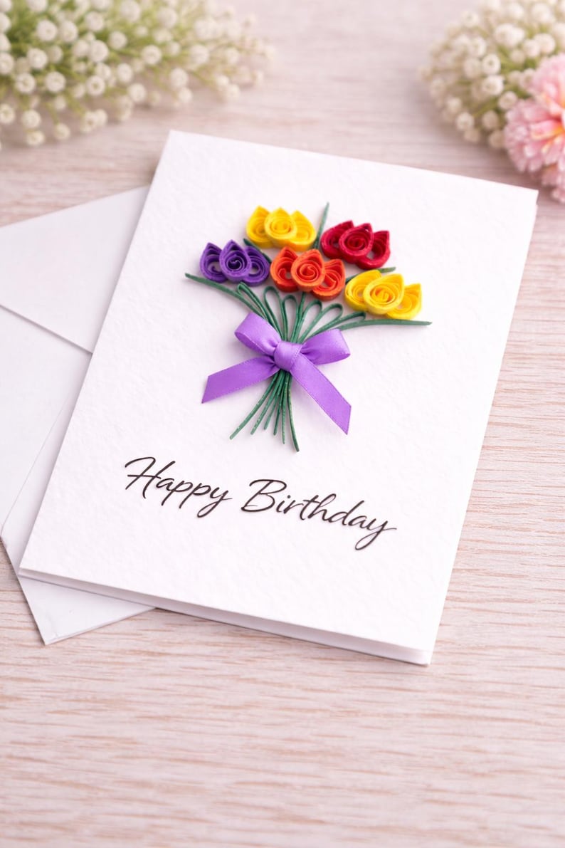 May include: A white birthday card featuring a paper flower bouquet in purple, yellow, red, and orange. The card is tied with a purple ribbon and has the words "Happy Birthday". An envelope is behind the card.