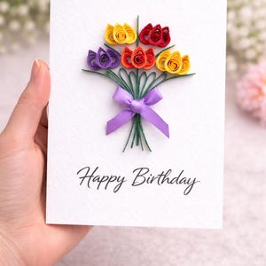 May include: A handmade birthday card featuring a paper flower bouquet. The flowers are yellow, red, orange, and purple, tied with a purple ribbon. The words "Happy Birthday" are written in black script.