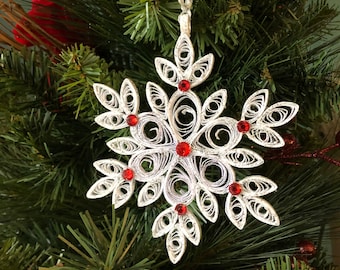 May include: A white paper snowflake ornament with red gem accents hangs from a silver ribbon against a backdrop of green Christmas tree branches. The snowflake is a decorative holiday item.