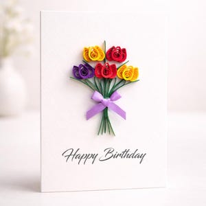 May include: A white birthday card featuring a quilled paper flower bouquet in yellow, red, and purple, tied with a purple ribbon. The words "Happy Birthday" are written in script.