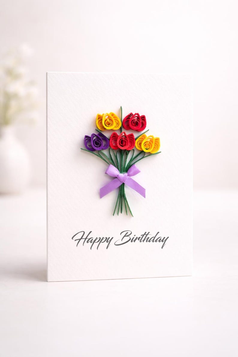 May include: A white birthday card featuring a quilled paper flower bouquet in yellow, red, and purple, tied with a purple ribbon. The words "Happy Birthday" are written in script.