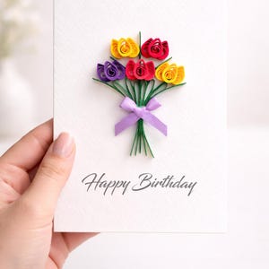 May include: A white birthday card featuring a paper flower bouquet in yellow, red, and purple, tied with a purple ribbon. The words "Happy Birthday" are printed in grey script below the flowers.