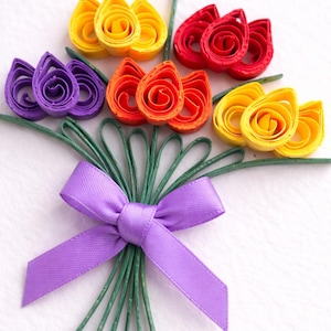 May include: A colourful paper flower bouquet made with quilling techniques. The flowers are in shades of yellow, red, orange, and purple, with green stems and a purple satin ribbon. The arrangement is a vibrant and decorative display.