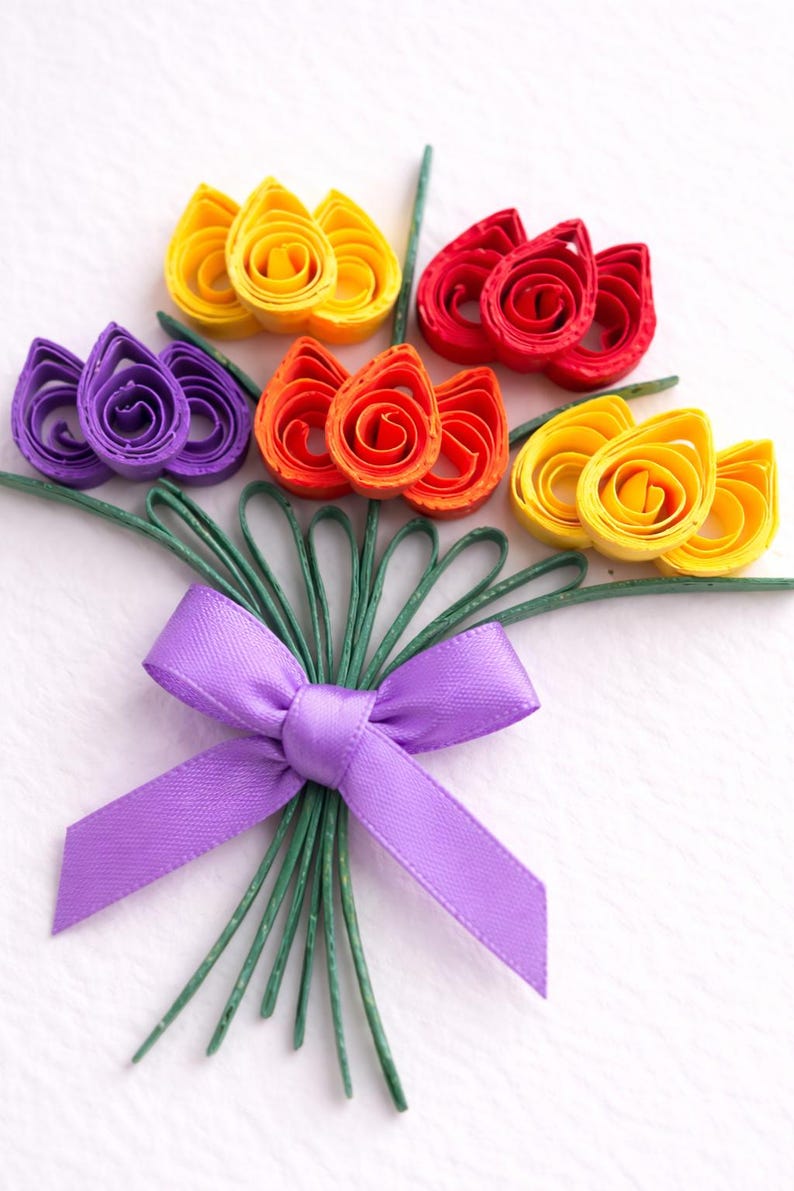 May include: A colourful paper flower bouquet made with quilling techniques. The flowers are in shades of yellow, red, orange, and purple, with green stems and a purple satin ribbon. The arrangement is a vibrant and decorative display.