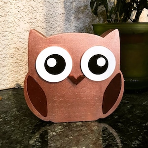 Könnte beinhalten: Eine hölzerne Eulenfigur mit großen, weißen Augen und einem braunen Körper. Die Eule ist in Kupferfarbe bemalt.
