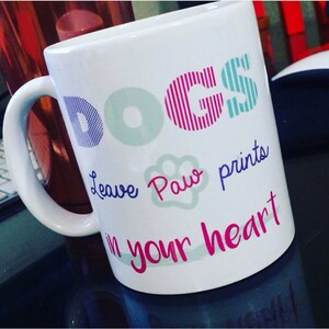 K&ouml;nnte beinhalten: Wei&szlig;e Keramiktasse mit dem Wort "DOGS" in bunten Buchstaben. Der Text "Leave paw prints in your heart" ist in lila und rosa Schrift geschrieben.