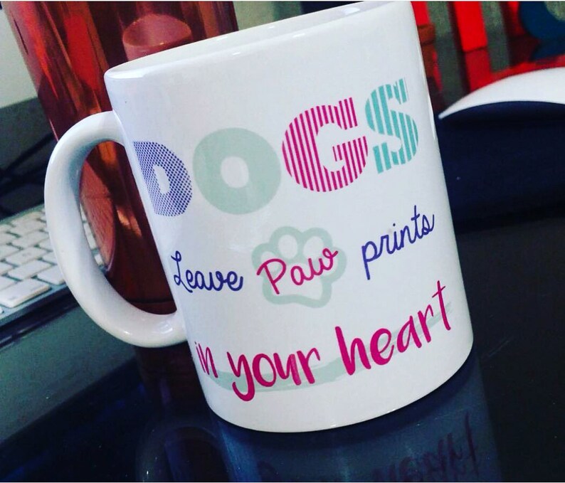 K&ouml;nnte beinhalten: Wei&szlig;e Keramiktasse mit dem Wort "DOGS" in bunten Buchstaben. Der Text "Leave paw prints in your heart" ist in lila und rosa Schrift geschrieben.