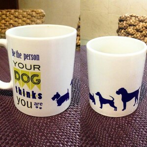 K&ouml;nnte beinhalten: Wei&szlig;e Keramiktasse mit blauem und gelbem Design. Die Tasse sagt "Sei die Person, die dein Hund denkt, dass du bist" mit Pfotenabdr&uuml;cken. Die Tasse hat eine blaue Silhouette eines Hundes an der Seite.