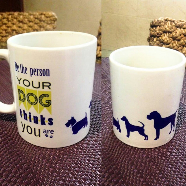 K&ouml;nnte beinhalten: Wei&szlig;e Keramiktasse mit blauem und gelbem Design. Die Tasse sagt "Sei die Person, die dein Hund denkt, dass du bist" mit Pfotenabdr&uuml;cken. Die Tasse hat eine blaue Silhouette eines Hundes an der Seite.