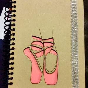 May include: A brown spiral bound notebook with a silver ribbon bookmark. The front cover features a pink cutout design of two ballet shoes.