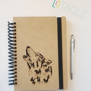 May include: A brown spiral bound notebook with a black elastic band closure. The cover features a laser-etched design of a wolf howling at the moon. The notebook is made of wood and has a rustic look.