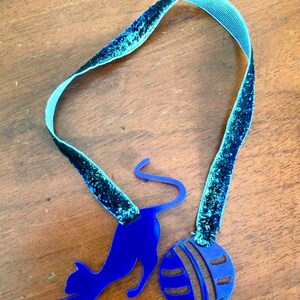 May include: A blue cat-shaped pendant with a ball charm, connected by a teal ribbon with glitter accents. The cat is in a playful pose, reaching towards the ball. The ribbon is approximately 12 inches long.