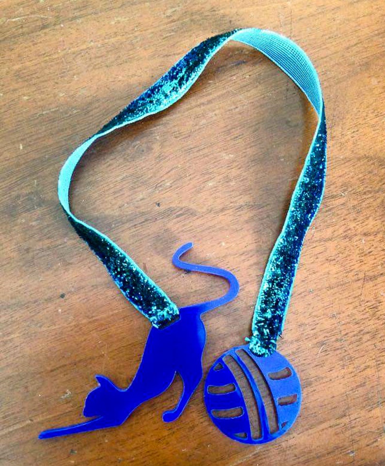May include: A blue cat-shaped pendant with a ball charm, connected by a teal ribbon with glitter accents. The cat is in a playful pose, reaching towards the ball. The ribbon is approximately 12 inches long.