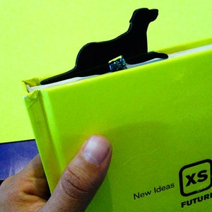 May include: A black dog-shaped bookmark is inserted into a bright green book. The book has the text "New Ideas" and "XS FUTURE" printed on it. The bookmark is made of a solid, dark material.