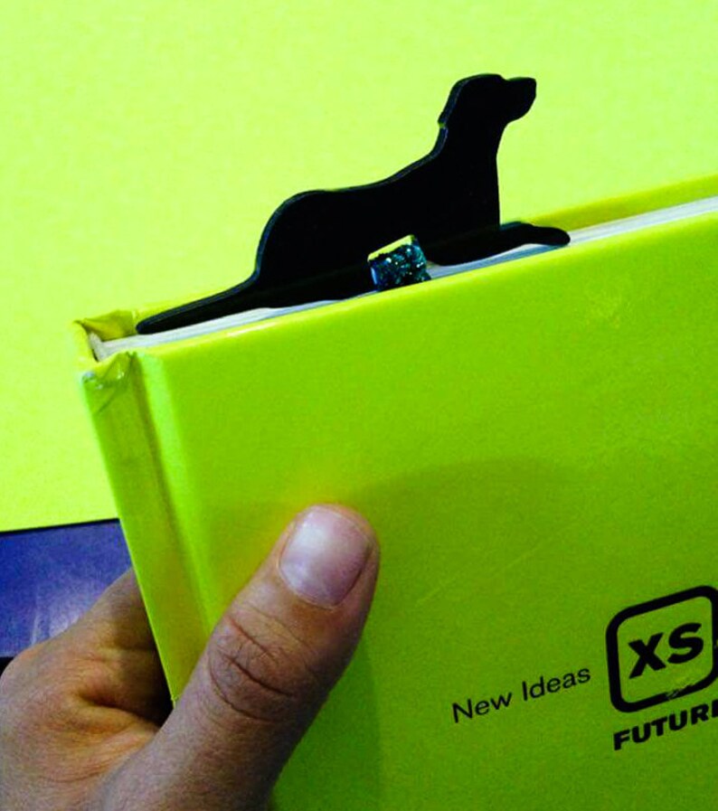 May include: A black dog-shaped bookmark is inserted into a bright green book. The book has the text "New Ideas" and "XS FUTURE" printed on it. The bookmark is made of a solid, dark material.