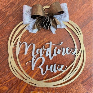 Könnte beinhalten: Ein goldfarbener Holz-Adventskranz mit dem Namen "Martinez Ruiz" in silbernen Lettern. Der Kranz ist mit einer weißen und goldenen Schleife und einer Tannenzapfen verziert.