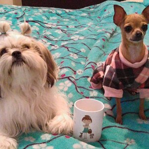 K&ouml;nnte beinhalten: Zwei Hunde, ein wei&szlig;er Shih Tzu mit Haarb&auml;ndern und ein brauner Chihuahua, der einen rosa-schwarzen karierten Pullover tr&auml;gt, sitzen auf einer t&uuml;rkisblauen Decke mit wei&szlig;em Blumenmuster. Eine wei&szlig;e Keramiktasse mit einem Cartoon-M&auml;dchen-Design steht im Vordergrund.