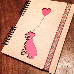 May include: A wooden journal with a pink silhouette of a dog holding a heart-shaped balloon. The journal has a gold glitter ribbon along the spine.