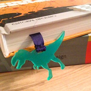 May include: A teal Tyrannosaurus Rex bookmark with a purple ribbon, marking a page in a book. The book has an orange and black cover with the text "EDITORIAL HIPERLIBRO". The bookmark is made of a translucent material.