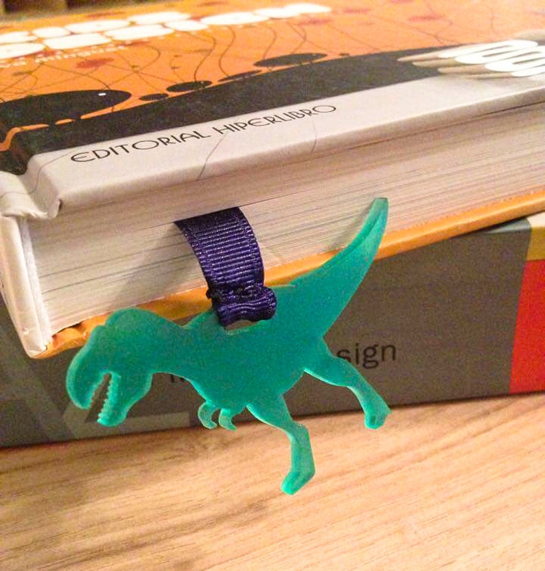 May include: A teal Tyrannosaurus Rex bookmark with a purple ribbon, marking a page in a book. The book has an orange and black cover with the text "EDITORIAL HIPERLIBRO". The bookmark is made of a translucent material.