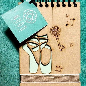 May include: A brown wooden notebook with a spiral binding. The cover features a laser-cut design of ballet shoes, a key, and hearts. The notebook has a light green label with the word "NUDO" and the text "Art and Design".