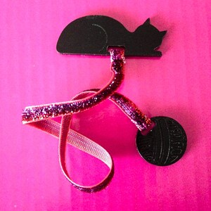 May include: A black cat-shaped hair accessory with a glittery pink ribbon and a round black accent. The accessory is set against a bright pink background, creating a playful contrast.