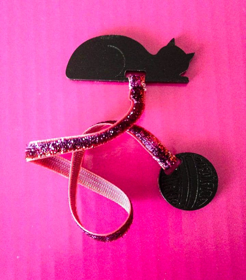 May include: A black cat-shaped hair accessory with a glittery pink ribbon and a round black accent. The accessory is set against a bright pink background, creating a playful contrast.