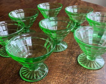 May include: A collection of eight green glass dessert dishes. Each dish has a pedestal base and a patterned bowl. The glass has a vibrant, translucent green color, and the dishes are arranged on a wooden surface.