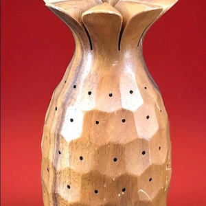 May include: A wooden pineapple-shaped vase with a textured surface and a flower-like top. The vase is light brown with dark brown accents and small black dot details. The background is a solid red.