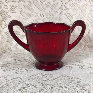 May include: A red glass creamer with two handles. The creamer has a scalloped rim and a footed base.