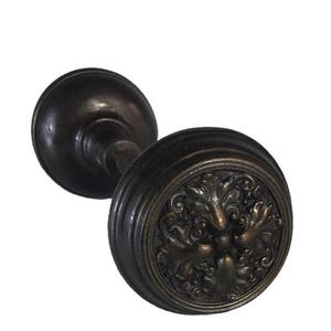 May include: Antique bronze door knob with a detailed floral design on the face. The knob has a round shape with a ridged edge and a stem connecting to a smaller, round base. The finish is a dark, aged bronze.
