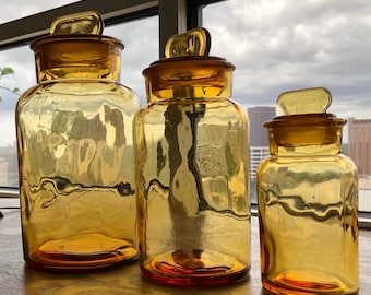 May include: Three amber glass apothecary jars with matching lids. The jars are different sizes, with a rectangular lid on top. The jars are sitting on a wooden surface, with a cityscape visible in the background.