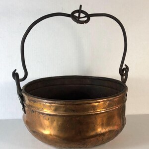 May include: A copper-colored metal cauldron with a dark, curved handle and a decorative loop at the top. The cauldron has a rounded body and a dark interior. The handle is attached to the cauldron with hooks.