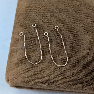 May include: Two delicate gold-coloured chain earring connectors with small loops at the top and a textured chain design. The chains are displayed on a brown, textured surface.