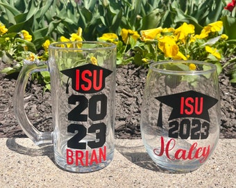 May include: Two glass beverage containers with personalized graduation designs. A clear glass mug with a black graduation cap and the text "ISU 2023 Brian" and a clear wine glass with a black graduation cap and the text "ISU 2023 Haley".